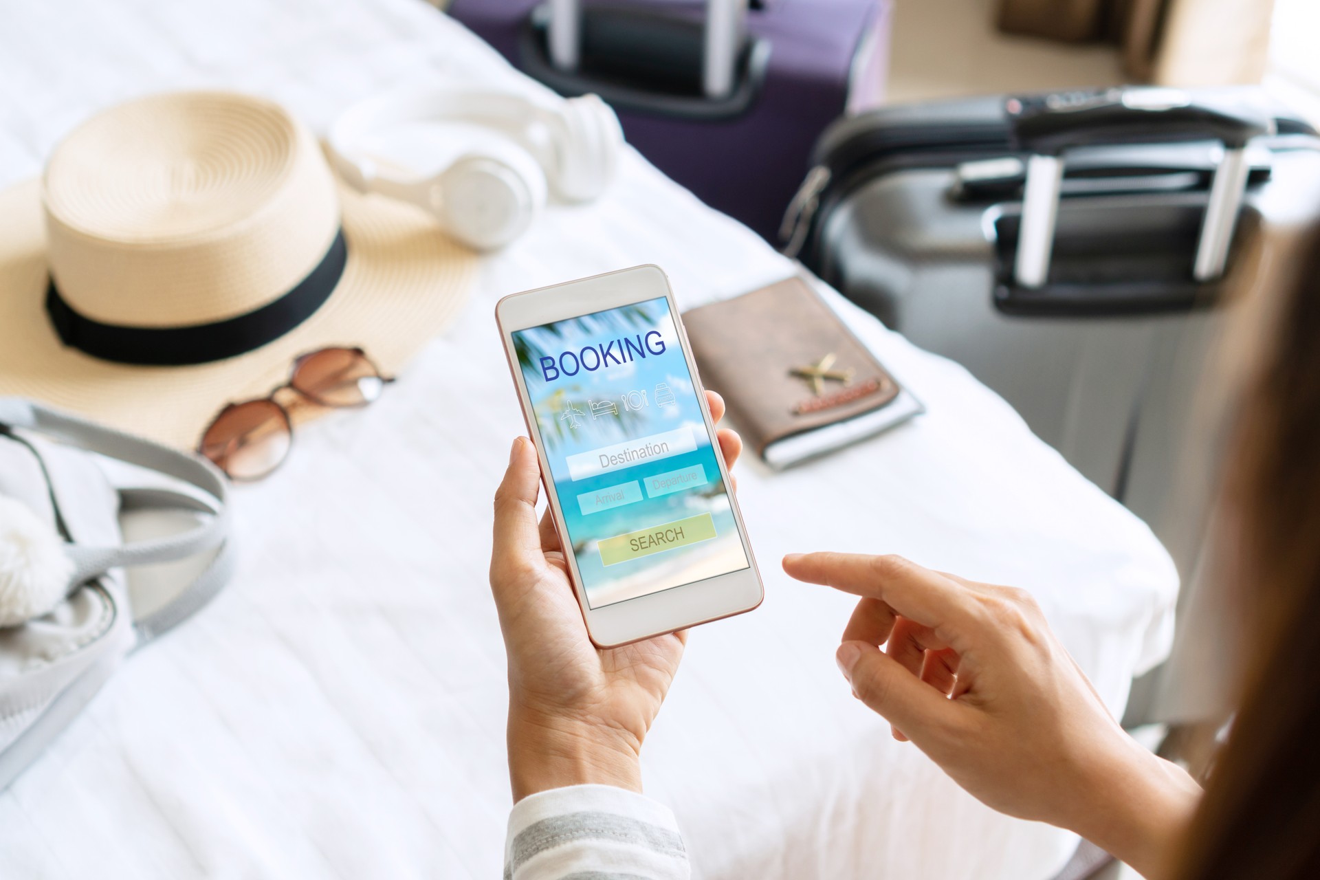 Woman hand holding smartphone with travel accessories on bed in hotel room. Travel, relaxation, journey, trip and vacation concepts. Top view  and copy space.